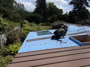 A Matrix Roofing roofer, tied off in a safety harness, installing a standing seam metal roof and skylight over blue self-adhered underlayment.
